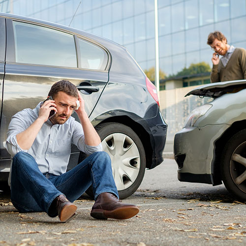 Ein Mann sitzt nach einem Unfall vor dem Auto und ruft an. Ein weiterer Mann steht hinter dem Auto und ruft ebenfalls