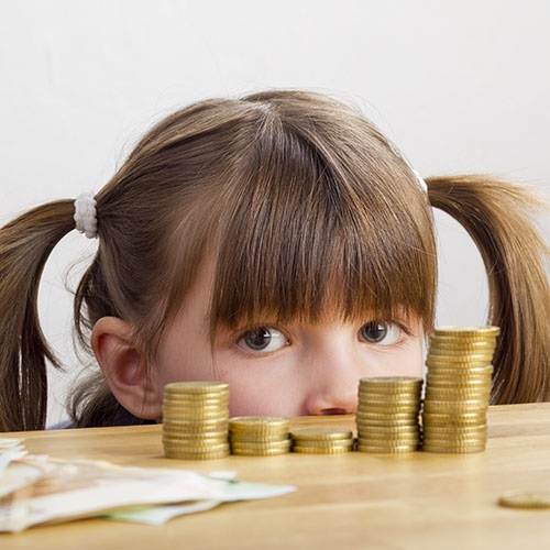 Mädchen schaut auf Geldtürme von Kindergeld Ein Mädchen mit Zöpfen schaut auf Geldtürme, die auf dem Tisch liegen von Kindergeld