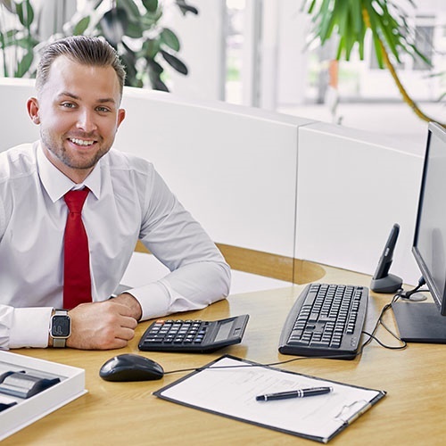 Ein lächelnder männlicher Serviceleiter sitzt am Tisch mit einem Computer, er ist selbstbewusst und professionell. Er trägt ein formelles Hemd und eine rote Krawatte.
