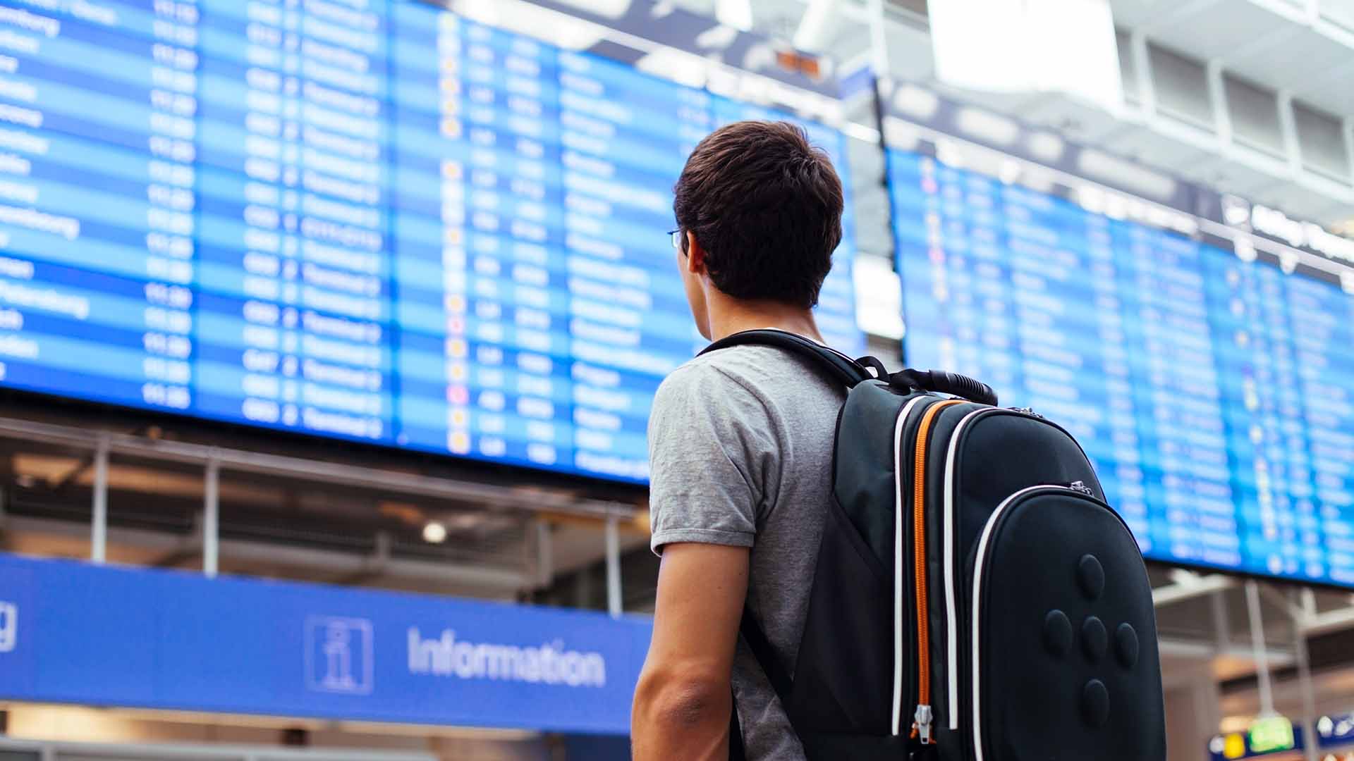 Ein Passagier mit Rucksack schaut auf eine Informationstafel im Flughafengebäude.